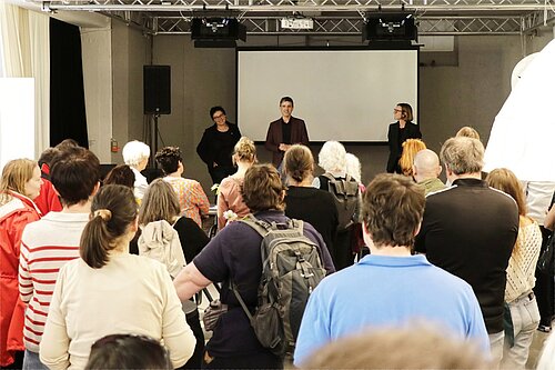 Ein Mann und zwei Frauen stehen auf der kleinen Bühne eines Veranstaltungssaals, vor ihnen stehen zahlreiche Zuhörerinnen und Zuhörer.